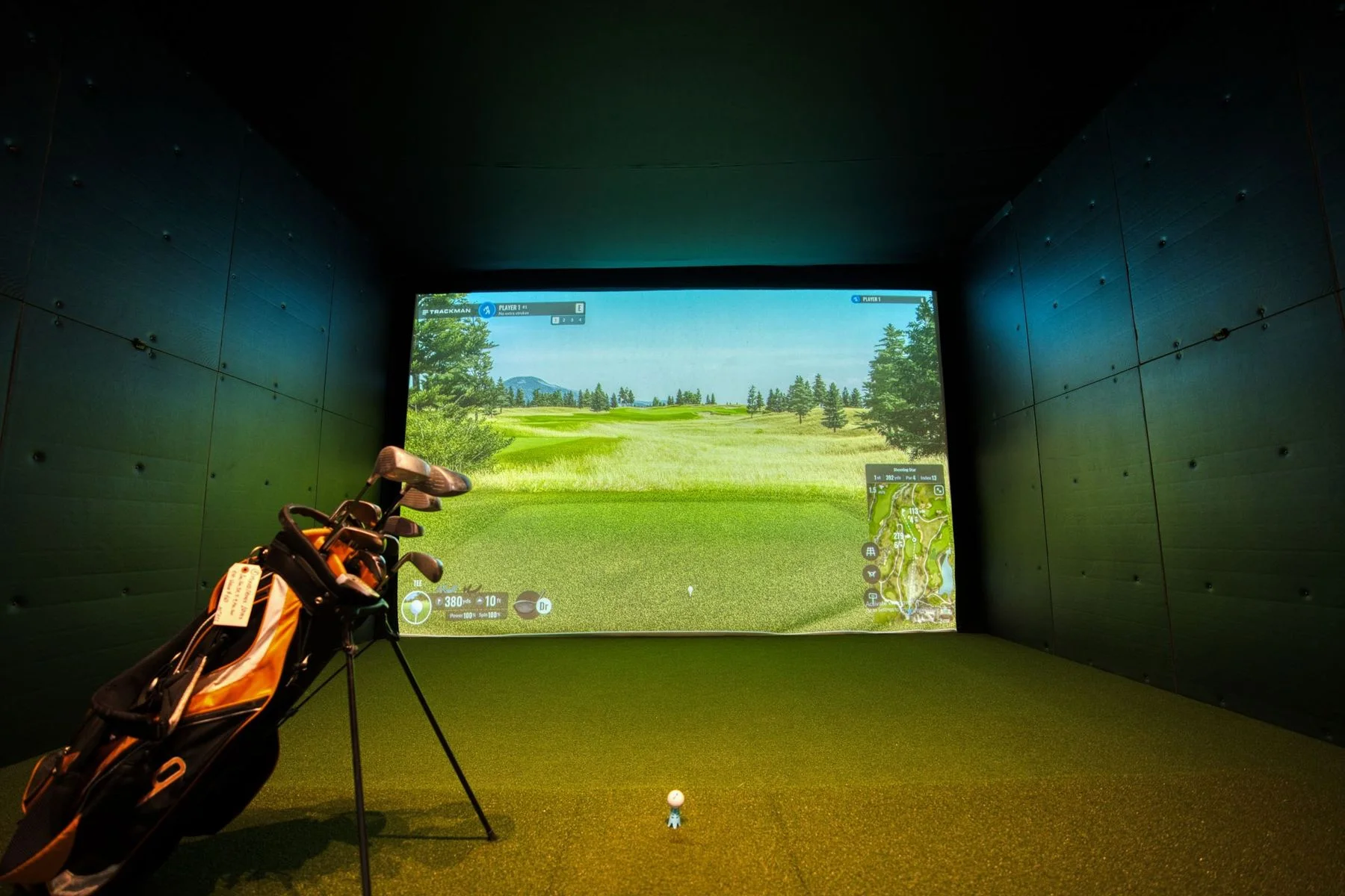 Real indoor golf simulator room with impact screen, hitting turf, golf bag, and projected course